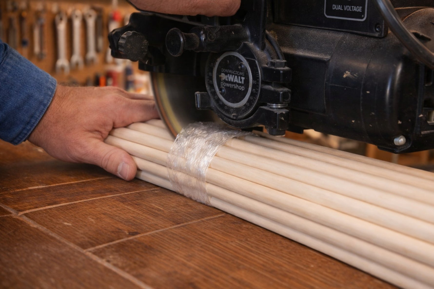 Radial arm saw cutting across a plastic-wrapped bundle of wooden dowels on a workshop bench while a worker steadies the dowels for a cross-cut.