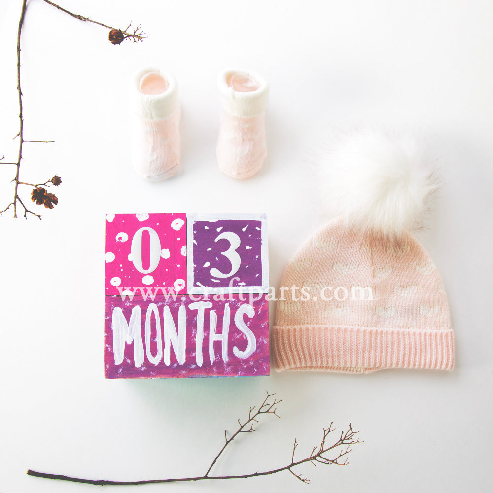 Hand-painted milestone block reading "0-3 Months," styled with baby socks and a hat for a photo prop